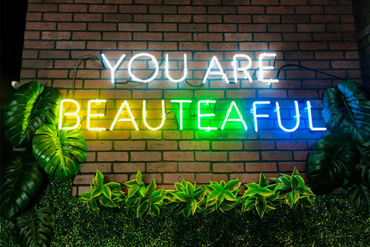 Neon sign saying "YOU ARE BEAUTEAFUL" in rainbow colors on brick wall with plants